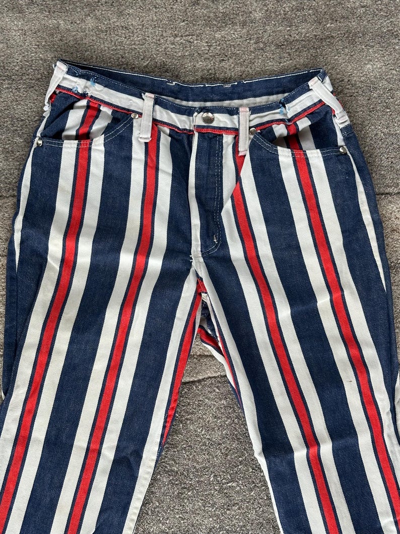 May include: Close-up of denim pants with vertical stripes in navy blue, white, and red. The pants have a classic five-pocket design with a button and zipper closure. The stripes run the length of the pants, creating a bold, eye-catching pattern.
