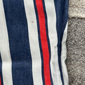 May include: Close-up of denim fabric with vertical stripes in navy blue, white, and red. The red stripe is centered between the white and navy blue stripes. The fabric has a textured appearance.