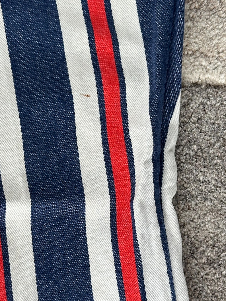 May include: Close-up of denim fabric with vertical stripes in navy blue, white, and red. The red stripe is centered between the white and navy blue stripes. The fabric has a textured appearance.