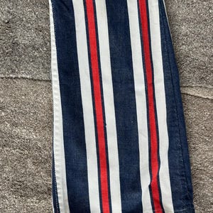 May include: Close-up of denim pants with vertical stripes. The design features alternating navy blue, white, and red stripes. The fabric is a medium weight denim, and the stripes run the length of the pants.