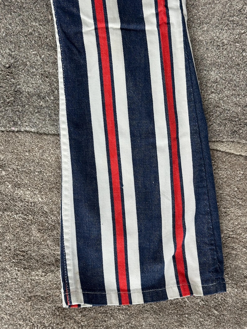 May include: Close-up of denim pants with vertical stripes. The design features alternating navy blue, white, and red stripes. The fabric is a medium weight denim, and the stripes run the length of the pants.