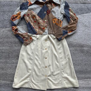 May include: A long-sleeved dress featuring a patchwork-style bodice with brown, white, and navy blue patterns including paisley and polka dots. The lower portion of the dress is a solid cream color with a vertical row of buttons down the center front.