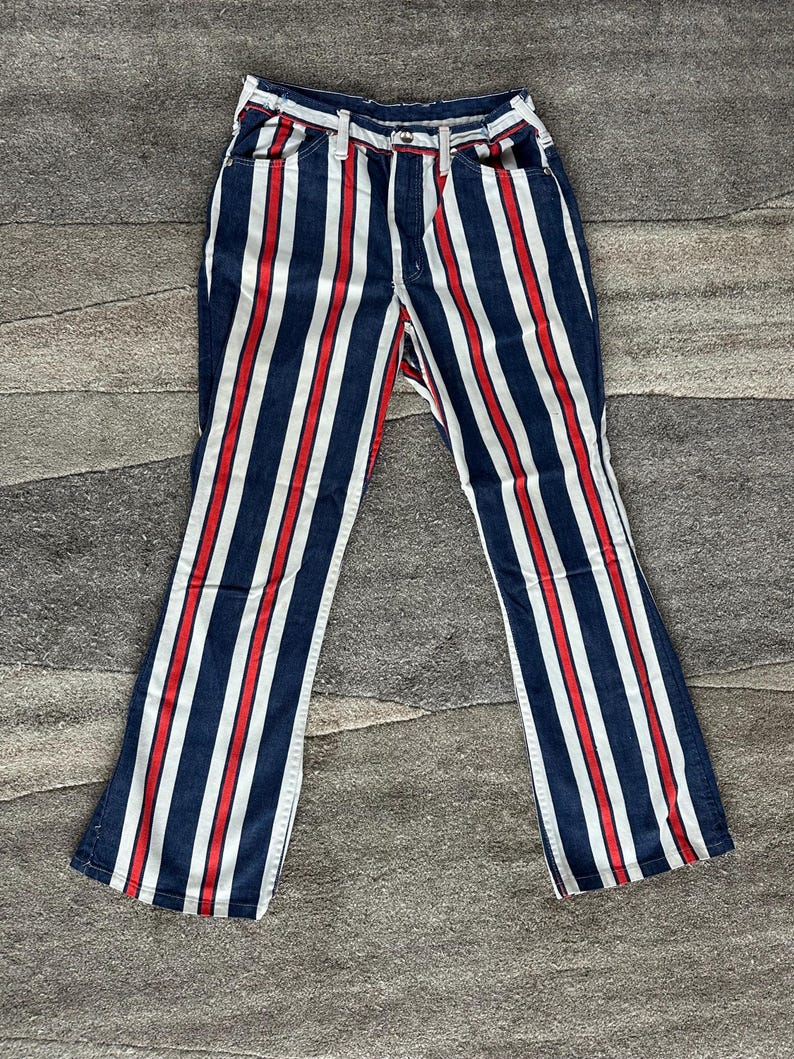 May include: Blue denim pants with vertical red and white stripes. The pants have a classic five-pocket design and a flared leg. The stripes are evenly spaced and run the full length of the pants.