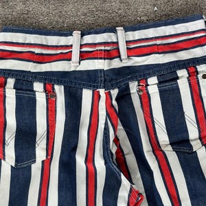 May include: Close-up of the back of a pair of denim jeans with a bold striped pattern. The stripes alternate between navy blue, white, and red. The jeans have two back pockets and a waistband with belt loops.