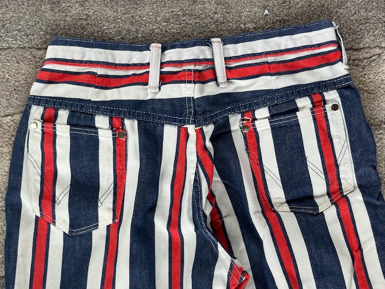 May include: Close-up of the back of a pair of denim jeans with a bold striped pattern. The stripes alternate between navy blue, white, and red. The jeans have two back pockets and a waistband with belt loops.