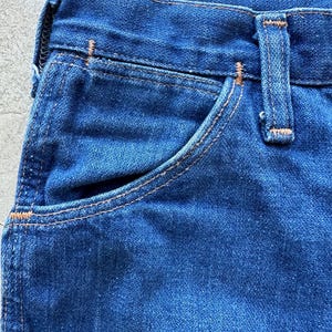 May include: Close-up of the top portion of a pair of blue denim jeans. The jeans have a light blue wash and a contrasting orange stitching. The image shows the top of the front pocket and the waistband.