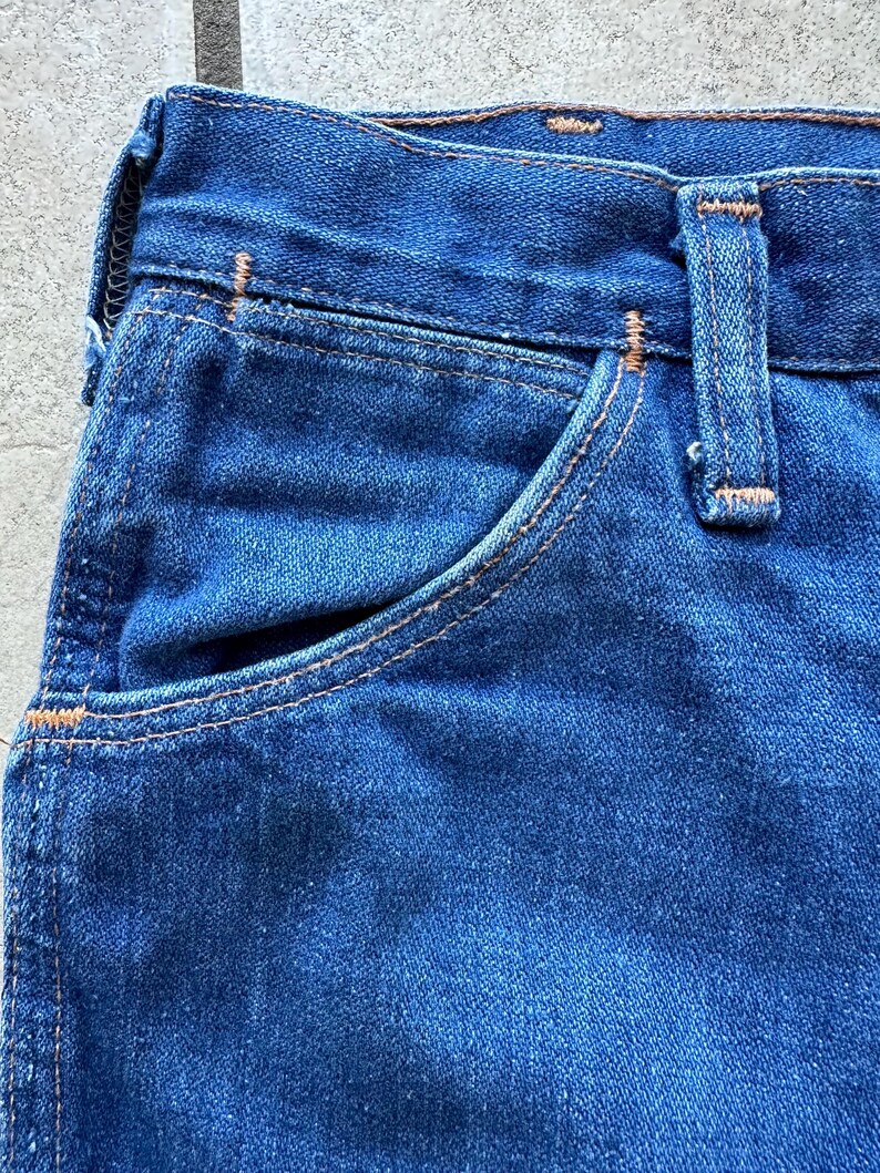 May include: Close-up of the top portion of a pair of blue denim jeans. The jeans have a light blue wash and a contrasting orange stitching. The image shows the top of the front pocket and the waistband.