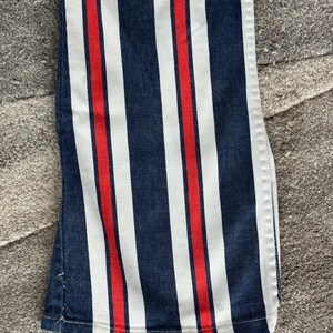 May include: Close-up of denim pants with vertical stripes in navy blue, white, and red. The fabric is a medium weight, and the stripes are evenly spaced. The pants have a classic, casual style.