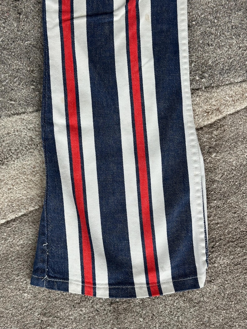 May include: Close-up of denim pants with vertical stripes in navy blue, white, and red. The fabric is a medium weight, and the stripes are evenly spaced. The pants have a classic, casual style.