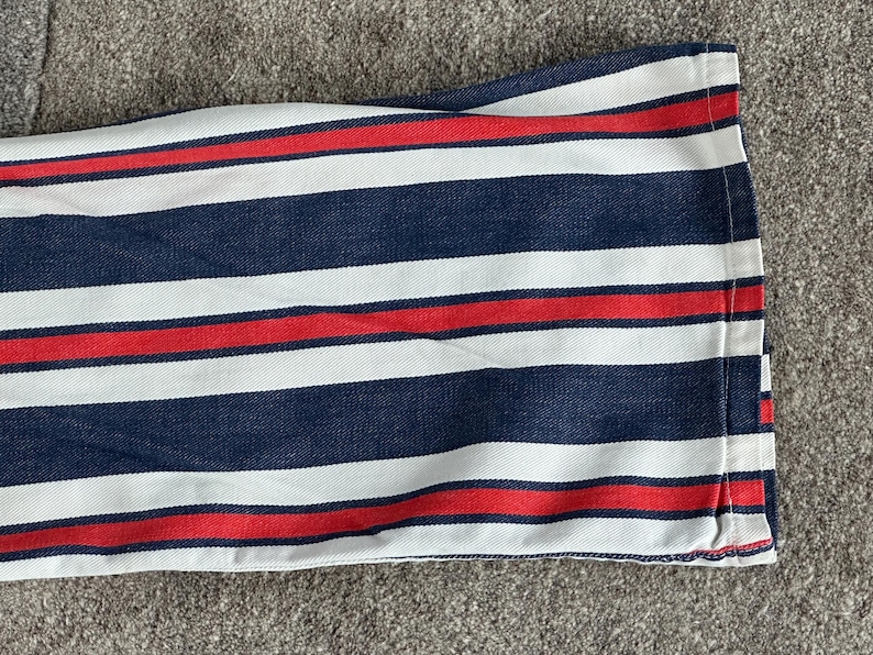 May include: Close-up of a fabric with a striped pattern. The design features alternating bands of navy blue, white, and red. The fabric is folded, revealing the edge with a red stitch.