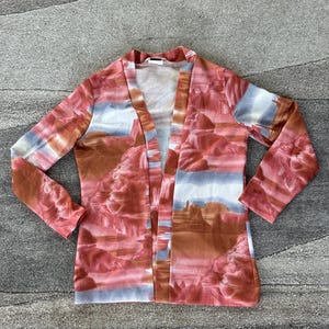 May include: A long-sleeved, open-front cardigan with a landscape print in shades of red, brown, and blue. The fabric features a canyon-like design, creating a unique and artistic look. The cardigan is laid flat on a textured surface.