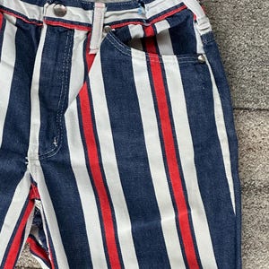 May include: Close-up of denim pants with vertical stripes in navy blue, white, and red. The pants have a button closure, belt loops, and pockets. The red stripe is centered between the white and navy blue stripes.