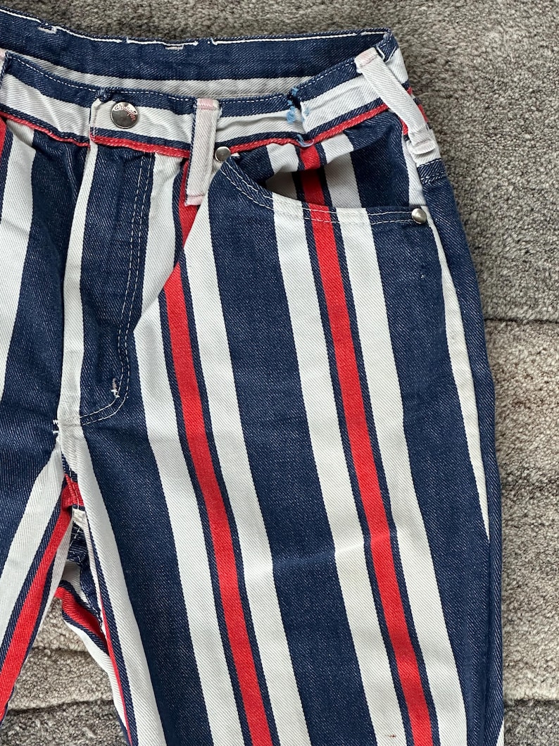 May include: Close-up of denim pants with vertical stripes in navy blue, white, and red. The pants have a button closure, belt loops, and pockets. The red stripe is centered between the white and navy blue stripes.