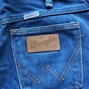 May include: A pair of blue denim jeans with a brown leather Wrangler label on the back pocket. The jeans have a classic five-pocket style and a straight leg.