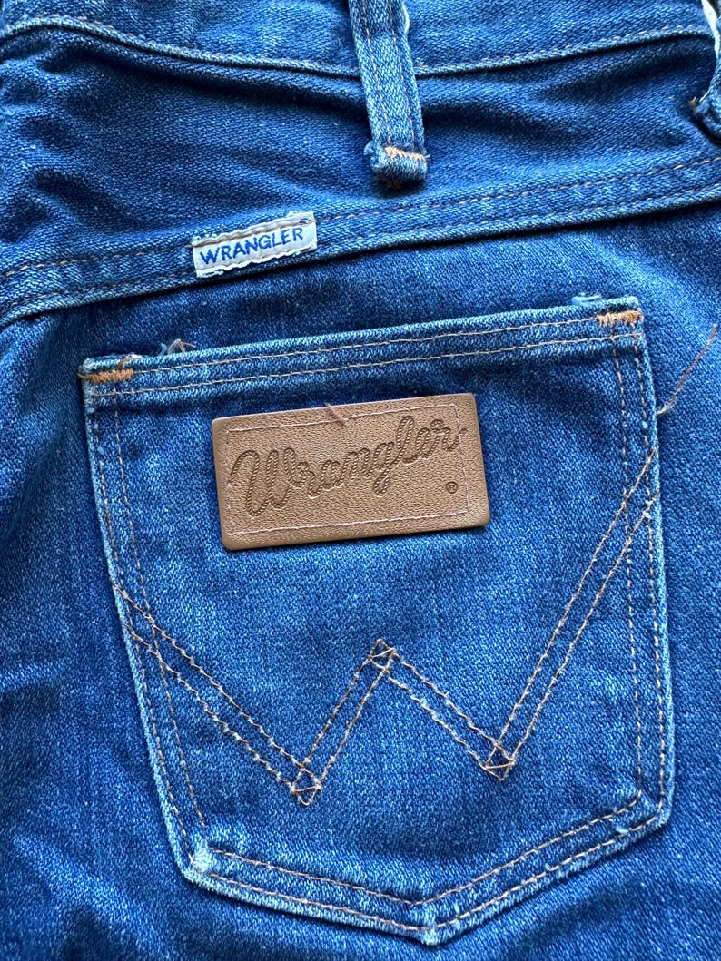 May include: A pair of blue denim jeans with a brown leather Wrangler label on the back pocket. The jeans have a classic five-pocket style and a straight leg.