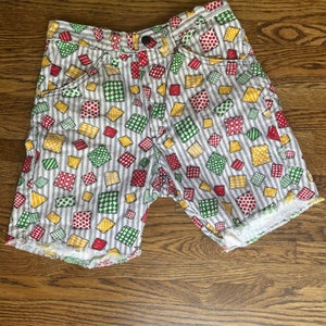 May include: White shorts with a colourful patchwork pattern of red, green, and yellow squares and dots. The shorts have a button closure and pockets, with a frayed hemline. The fabric has vertical blue pinstripes.