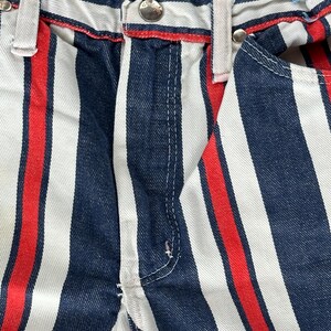 May include: Close-up of vertically striped denim pants. The pants feature alternating wide stripes of white, navy blue, and red. The waistband has a red trim and silver button. The overall style is casual and eye-catching.