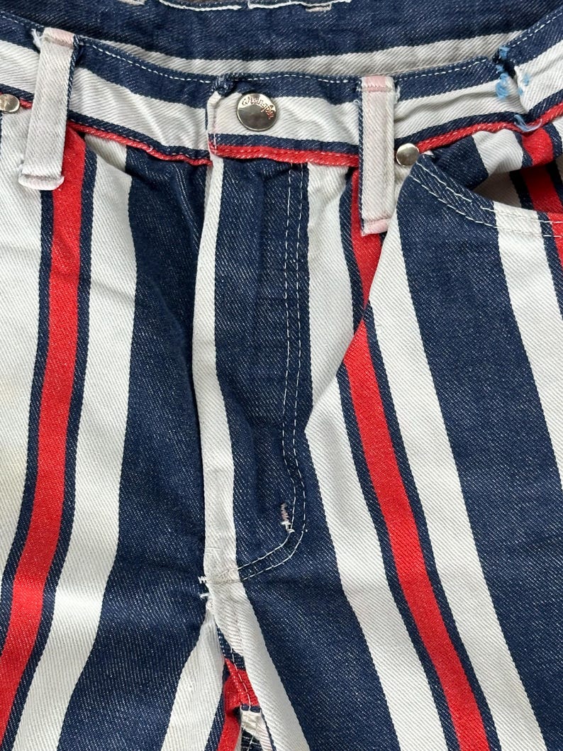 May include: Close-up of vertically striped denim pants. The pants feature alternating wide stripes of white, navy blue, and red. The waistband has a red trim and silver button. The overall style is casual and eye-catching.