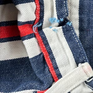 May include: Close-up of a section of striped fabric. The fabric features alternating white and navy blue vertical stripes, with a red stripe accent. The edges are visible, showing the texture and construction of the material.