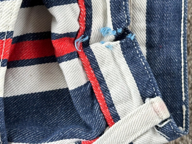 May include: Close-up of a section of striped fabric. The fabric features alternating white and navy blue vertical stripes, with a red stripe accent. The edges are visible, showing the texture and construction of the material.