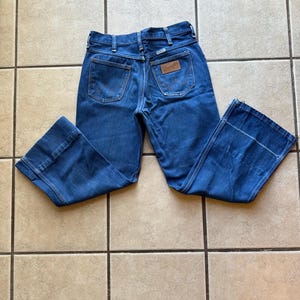 May include: A pair of blue denim jeans with a wide leg and a high waist. The jeans have a brown leather patch on the back with the Wrangler logo.