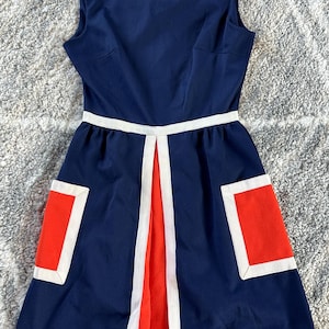 May include: A sleeveless navy blue dress featuring a high neckline, a white waistband, and two rectangular orange pockets with white trim. The skirt includes a central orange panel framed by white vertical stripes and a white hemline border.