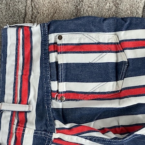May include: Close-up of a section of striped denim fabric. The fabric features alternating red, white, and navy blue stripes. A pocket and a small button are visible. The fabric appears to be part of a pair of shorts or pants.