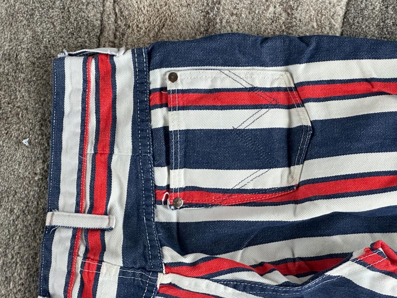 May include: Close-up of a section of striped denim fabric. The fabric features alternating red, white, and navy blue stripes. A pocket and a small button are visible. The fabric appears to be part of a pair of shorts or pants.
