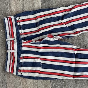 May include: Blue denim pants with horizontal red and white stripes. The pants have a classic five-pocket design, belt loops, and a button and zipper closure. The stripes are evenly spaced across the fabric.