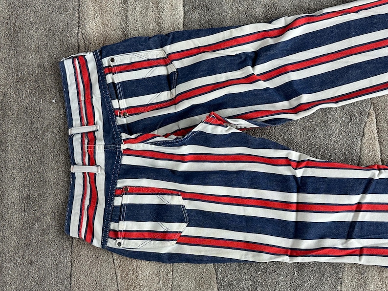 May include: Blue denim pants with horizontal red and white stripes. The pants have a classic five-pocket design, belt loops, and a button and zipper closure. The stripes are evenly spaced across the fabric.