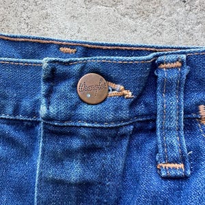 May include: Close-up of the waistband of a pair of blue denim jeans with a copper-colored Wrangler button.