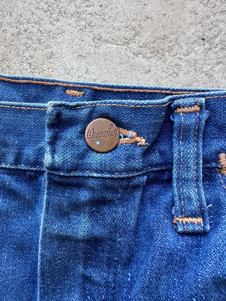 May include: Close-up of the waistband of a pair of blue denim jeans with a copper-colored Wrangler button.