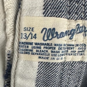May include: Close-up of a tag from a Wrangler garment. The tag is cream-colored with black text, displaying the size "13/14" and the Wrangler logo. The fabric is a black and white checkered pattern.