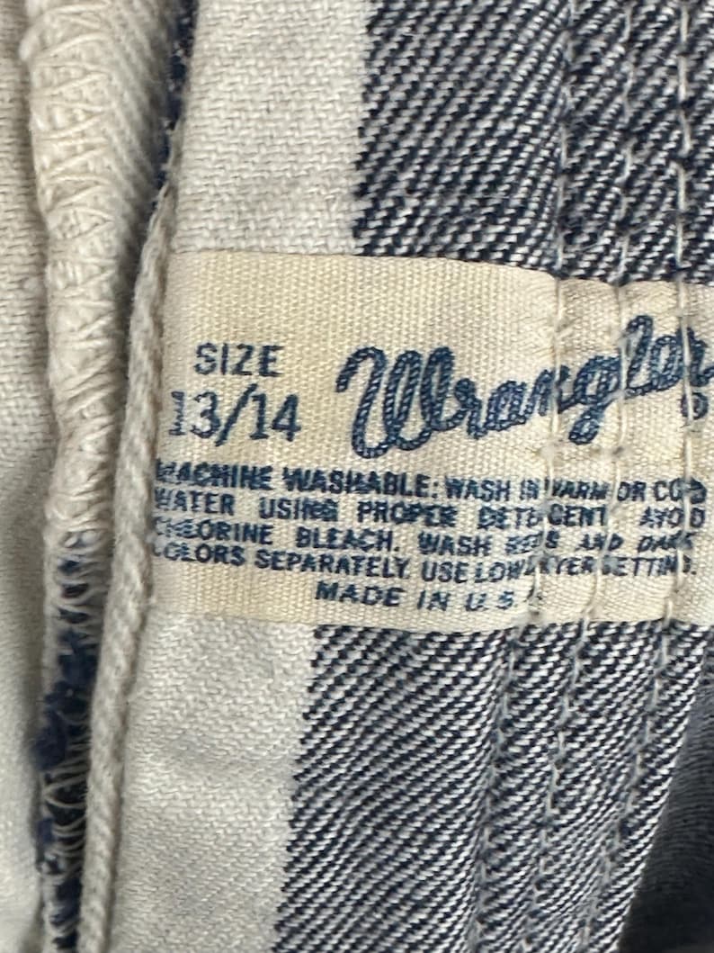 May include: Close-up of a tag from a Wrangler garment. The tag is cream-colored with black text, displaying the size "13/14" and the Wrangler logo. The fabric is a black and white checkered pattern.