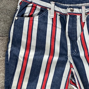 May include: Close-up of denim pants with vertical stripes in navy blue, white, and red. The pants have a classic five-pocket design, belt loops, and a button closure. The stripes run the length of the pants.