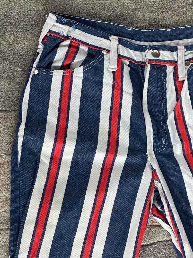 May include: Close-up of denim pants with vertical stripes in navy blue, white, and red. The pants have a classic five-pocket design, belt loops, and a button closure. The stripes run the length of the pants.