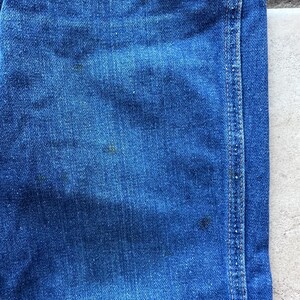 May include: Close-up of blue denim fabric with a single seam running down the right side. The fabric has a textured, woven appearance.