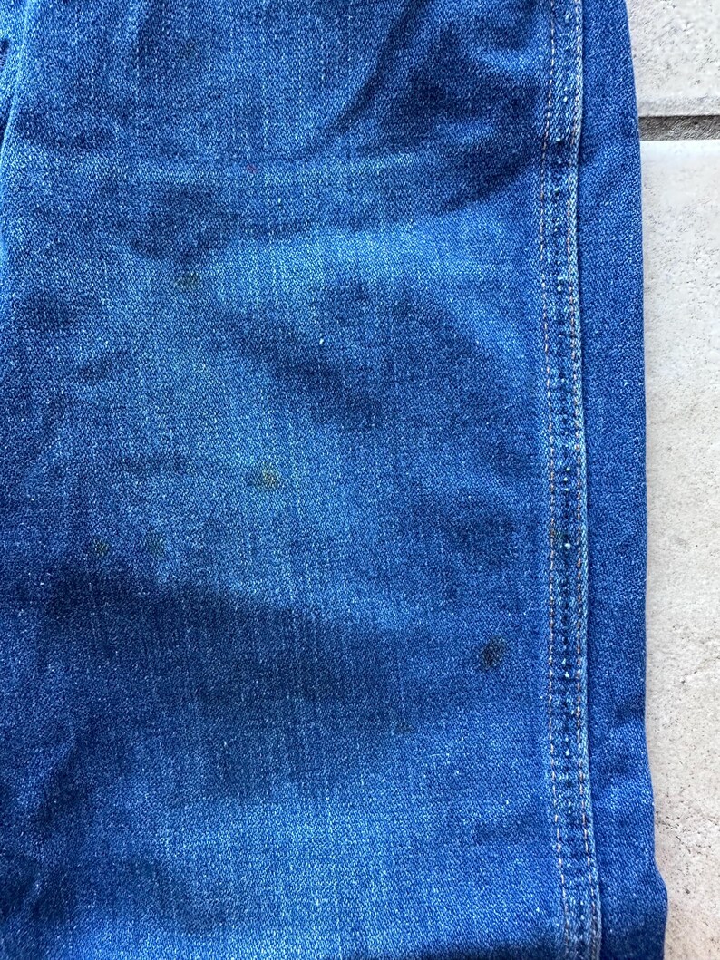 May include: Close-up of blue denim fabric with a single seam running down the right side. The fabric has a textured, woven appearance.