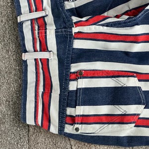May include: Close-up of a section of striped fabric in red, white, and navy blue. The fabric appears to be denim or a similar material, with a pocket and belt loops visible. The stripes are horizontal and evenly spaced.