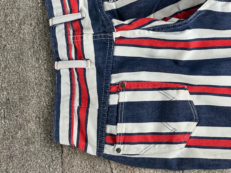 May include: Close-up of a section of striped fabric in red, white, and navy blue. The fabric appears to be denim or a similar material, with a pocket and belt loops visible. The stripes are horizontal and evenly spaced.