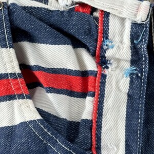 May include: Close-up of a section of striped fabric. The fabric features alternating bands of navy blue, white, and red. White stitching is visible along the seams, and a silver button is present. The fabric appears to be denim.