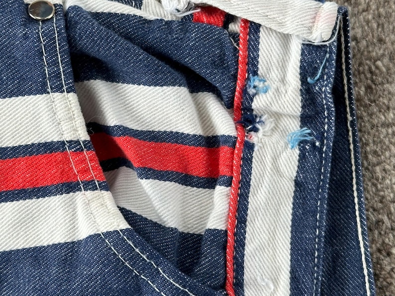 May include: Close-up of a section of striped fabric. The fabric features alternating bands of navy blue, white, and red. White stitching is visible along the seams, and a silver button is present. The fabric appears to be denim.