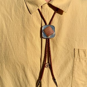 May include: A bolo tie featuring a square silver-toned metal slide with a central diamond-shaped peach-colored stone. The slide is attached to a braided brown leather cord that terminates in two pointed silver-toned metal tips at the bottom ends.