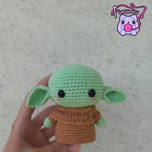 Puede incluir: Mu&ntilde;eco de ganchillo de un personaje con cabeza grande, orejas grandes y ojos negros. El mu&ntilde;eco es principalmente verde claro con un atuendo marr&oacute;n. El mu&ntilde;eco se sostiene en una mano. El logo "orgumiyu" est&aacute; en la esquina superior derecha.