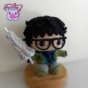 May include: A crocheted figure of a person with black hair, wearing glasses, a green jacket, and gray pants. The figure is holding a gray crocheted object in their right hand. The figure is sitting on a wooden base.
