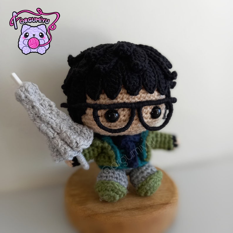 May include: A crocheted figure of a person with black hair, wearing glasses, a green jacket, and gray pants. The figure is holding a gray crocheted object in their right hand. The figure is sitting on a wooden base.