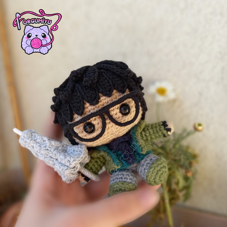 May include: A crocheted amigurumi figure of a person wearing glasses, a green jacket, and blue jeans. The figure is holding a gray crocheted umbrella.
