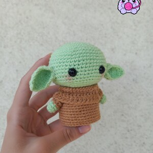 Puede incluir: Una mu&ntilde;eca de ganchillo de un personaje con orejas grandes, piel verde y un atuendo marr&oacute;n. La mu&ntilde;eca tiene ojos negros y un ligero rubor rosado. La mu&ntilde;eca se sostiene en una mano. La imagen tambi&eacute;n incluye un logotipo con el texto "Forgumiyu".