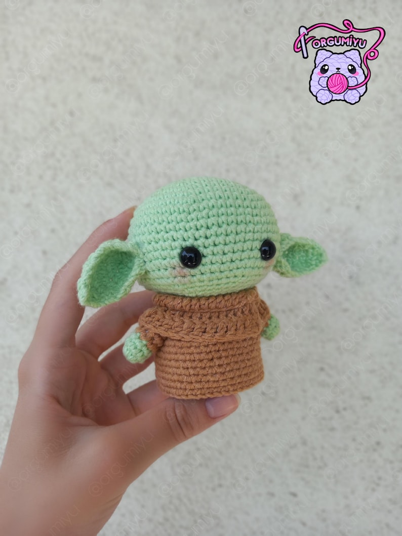 Puede incluir: Una mu&ntilde;eca de ganchillo de un personaje con orejas grandes, piel verde y un atuendo marr&oacute;n. La mu&ntilde;eca tiene ojos negros y un ligero rubor rosado. La mu&ntilde;eca se sostiene en una mano. La imagen tambi&eacute;n incluye un logotipo con el texto "Forgumiyu".