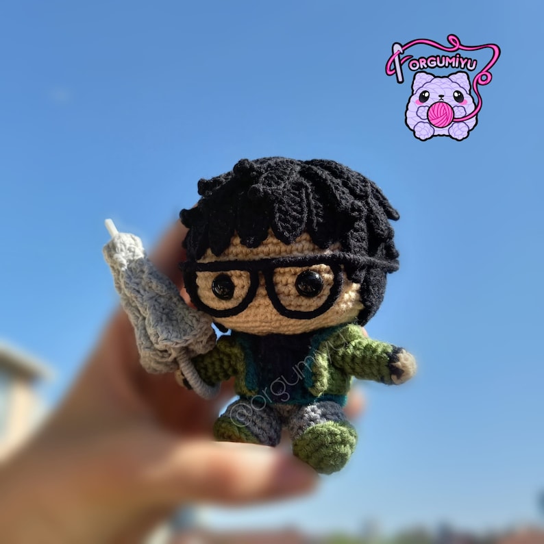 May include: A crocheted plush toy of a person wearing glasses and a green jacket, holding a gray crocheted object. The toy is against a blue sky background.